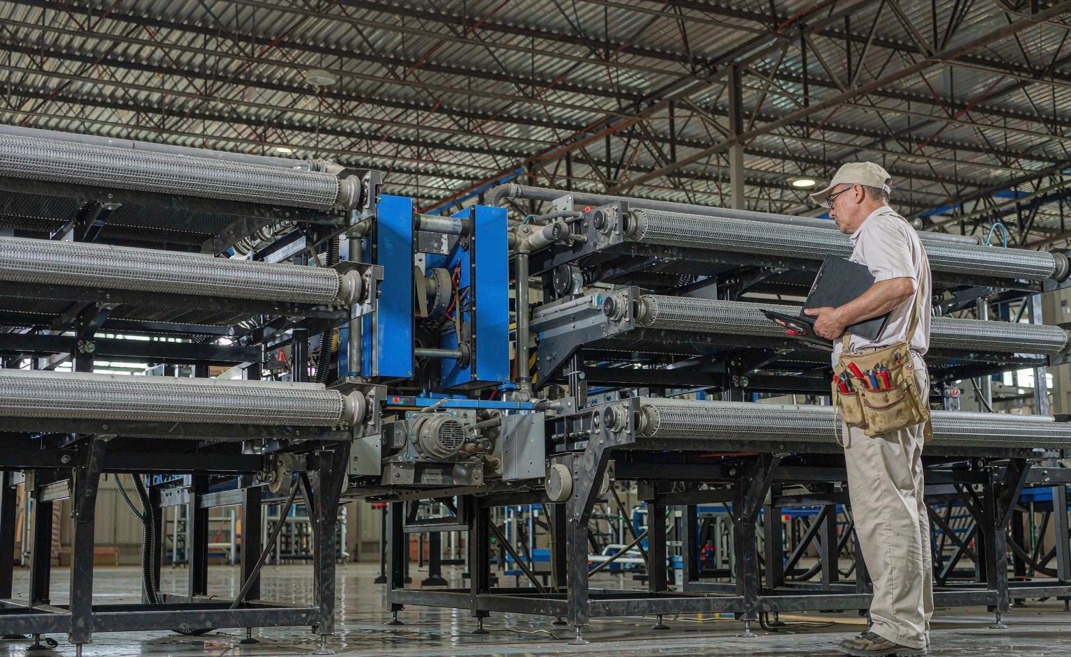 Maintenance technician inspecting a cross-belt sorter conveyor system with clipboard in a logistics warehouse
